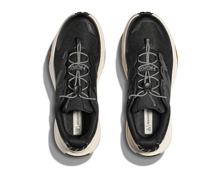 Top view of HOKA Mens Transport Wide Black/White sneakers with white soles, unique patterned laces, and a VIBRAM outsole for superior traction, displayed side by side on a plain background.
