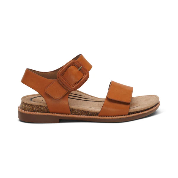 The Aetrex Women's Claire Cognac by AETREX WORLDWide INC is a brown leather sandal with a flat sole, open toe, front and ankle straps with a large buckle, cushioned footbed, cork accent, and excellent arch support.