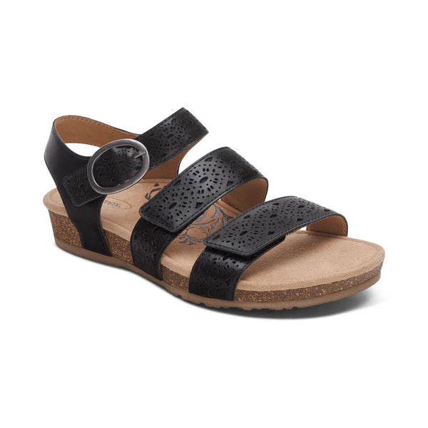 The Aetrex Womens Lilly Cutout Black sandal by AETREX WORLDWide INC features three perforated leather straps, a side buckle, black open-toe design, low cork wedge heel, and a cushioned tan footbed with arch support and a subtle floral pattern.
