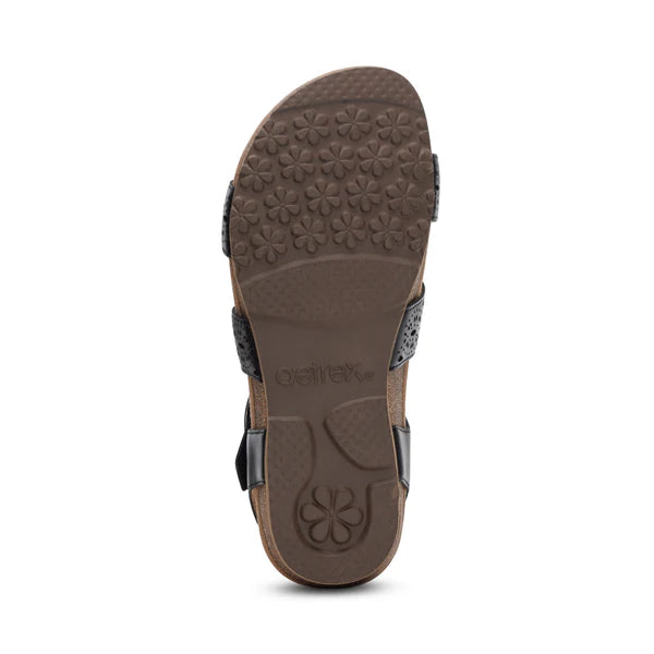 The image shows the sole of the Aetrex Womens Lilly Cutout Black sandal by AETREX WORLDWide INC, featuring a floral pattern, two black side straps, and Aetrex with a flower logo embossed near the arch for signature support.