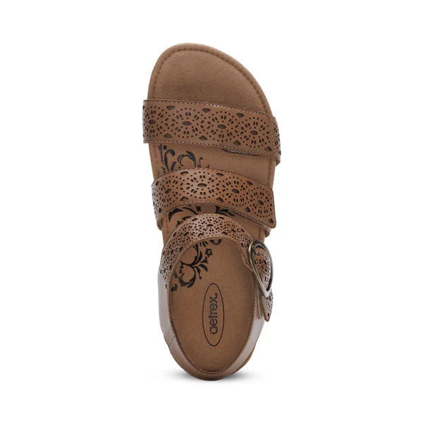A taupe sandal with three decorative straps, floral cutout patterns, a large side buckle, and a cushioned memory foam footbed for comfort. Seen from above, the Aetrex Womens Lilly Cutout Taupe by AETREX WORLDWide INC offers arch support.