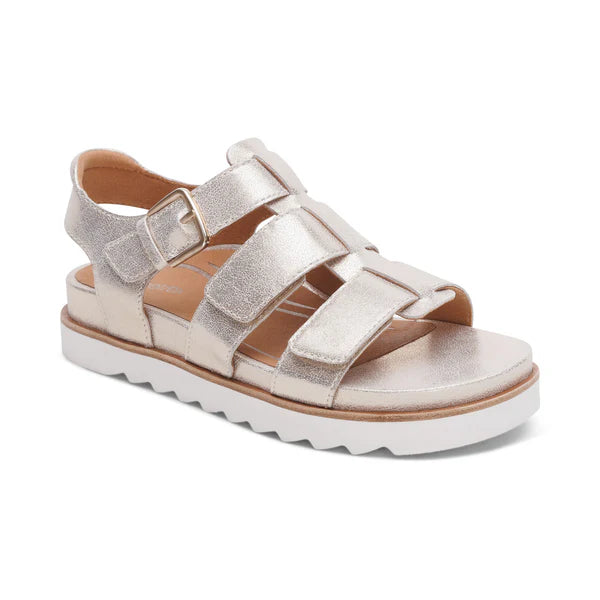 The Aetrex Women's Romy Champagne sandal by AETREX WORLDWide INC features metallic gold straps, an adjustable buckle, cushioned footbed for foot pain relief, and a thick white ridged sole, shown angled on a white background.