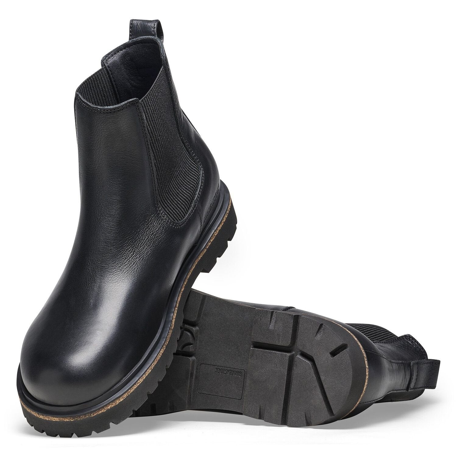 The BIRKENSTOCK Mens Highwood Slip On Black is a pair of black natural leather Chelsea boots with chunky rubber soles, side elastic panels, visible tread pattern, and Deep Blue Footbed comfort. One boot stands upright; the other lies on its side.