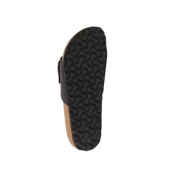 The image shows the bottom view of the BIRKENSTOCK Womens Madrid Big Buckle Black sandal, featuring a black, textured rubber sole with a wavy geometric tread, cork footbed, and black oiled nubuck leather upper straps.
