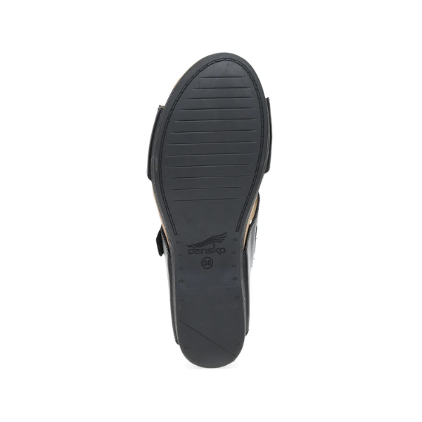 The image shows the black rubber sole of the Dansko Womens Shannon Black Burnished wedge slide sandal, viewed from below. It features horizontal grooves, a raised heel, and an embossed DANSKO logo for added foot pain relief.