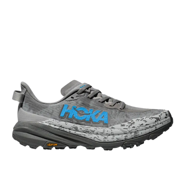 The HOKA Womens Speedgoat 6 Stellar Grey/Asteroid Wide features a gray upper, blue HOKA logo, black laces, and a rugged black-and-gray sole with the yellow Vibram Megagrip logo—ideal for trail running.