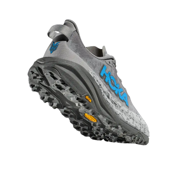 The HOKA Womens Speedgoat 6 Stellar Grey/Asteroid Wide, shown in gray with rugged black and gray Vibram Megagrip tread, a blue HOKA logo on the side, and a yellow Vibram logo on the sole, is displayed from a rear angle on a white background.