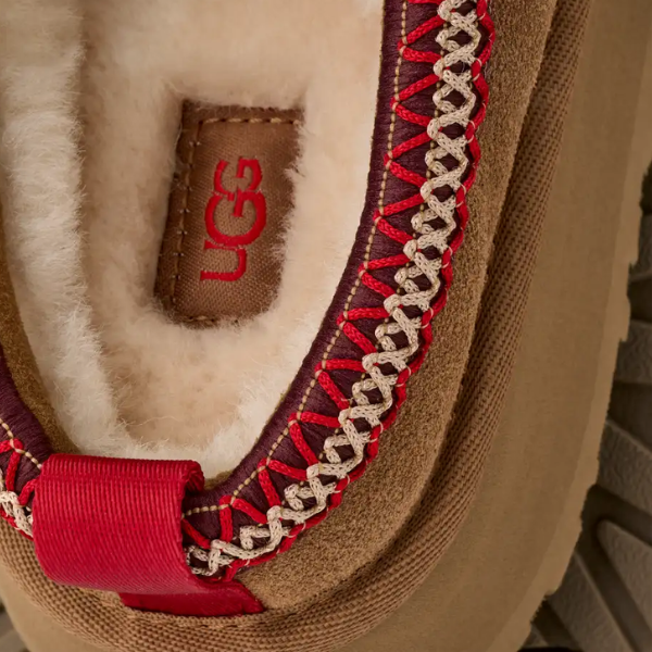 Close-up of the inside heel of an UGG Women's Tazz II Chestnut shoe, showing white sheepskin lining, red and beige stitching, a red UGG logo on a brown tag, and a red fabric pull tab.