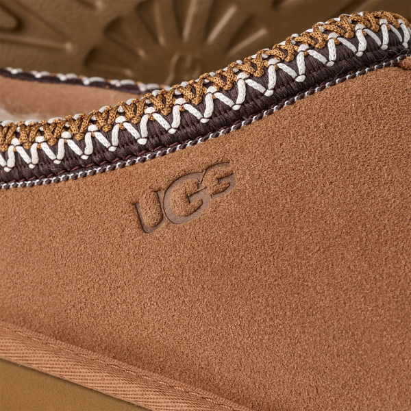 Close-up of the UGG Men's Tasman II Chestnut featuring a tan suede upper, embossed UGG logo, and detailed brown, white, and tan stitching along the edge.