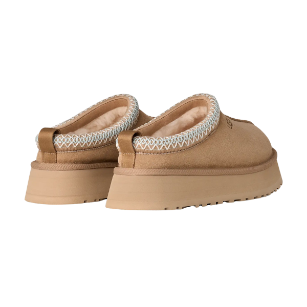 The UGG Women's Tazz II Sand slip-ons feature tan suede, thick textured soles, cozy sheepskin lining, and light blue and white decorative stitching at the opening. Shown from a back angled view. Brand: UGG.