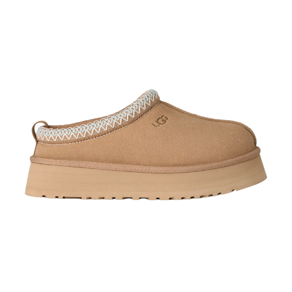 The UGG Women’s Tazz II Sand slipper features tan suede, a sheepskin lining, thick ridged sole, and decorative ankle stitching, displayed in a side profile on a white background.