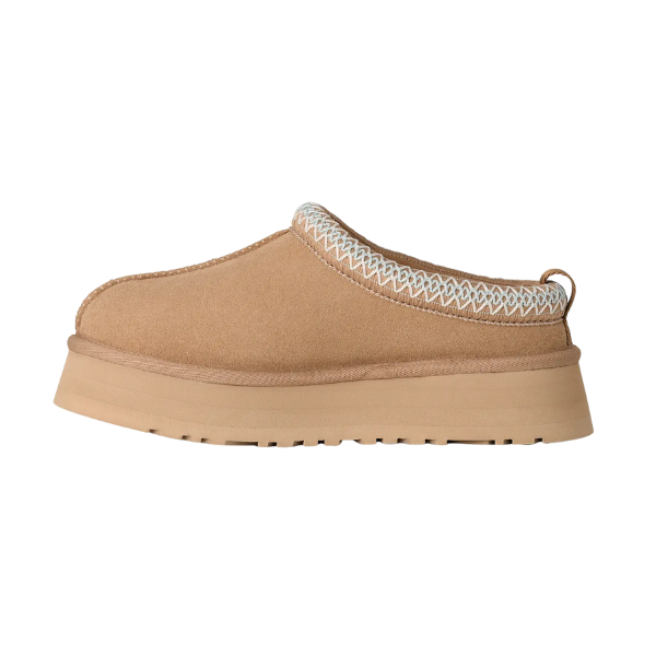 The UGG Women's Tazz II Sand slip-on, shown in side profile on a white background, features a tan color, thick textured sole, white decorative collar stitching, and plush sheepskin lining.