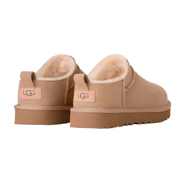 The UGG Women's Classic Micro Sand slippers feature tan suede, plush cream lining, and durable rubber soles, blending classic sheepskin boot comfort with slip-on ease. Shown from the back and side against a white background.