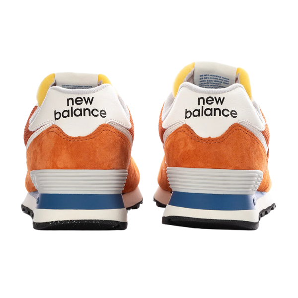 A pair of New Balance Unisexs 574 Infield Clay/Sea Salt shoes is shown from the back, with the NEW BALANCE name on the heel and blue and gray accents on the soles.