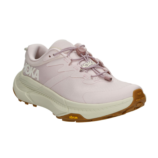 The HOKA Womens Transport in Cosmic Pearl/Oat Milk/Pink features a light pink upper, white logo, beige midsole, and brown Vibram sole. Designed for hiking, its shown at a front diagonal angle on a white background.