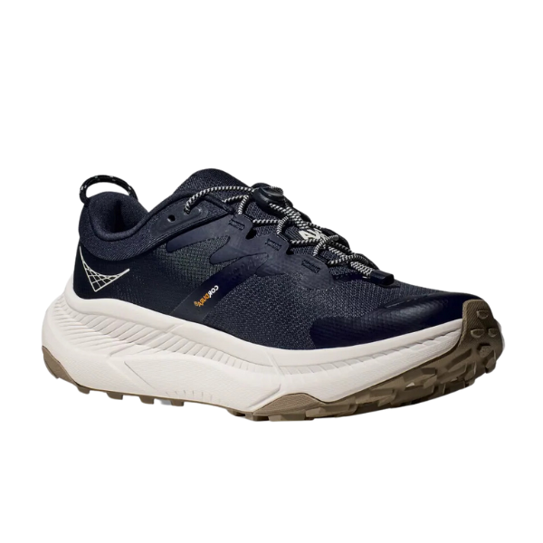 The HOKA Mens Transport Wide Navy is a dark blue vegan athletic shoe with white and gray laces, a white cushioned sole, and a brown VIBRAM outsole for trail or running, shown against a plain white background.