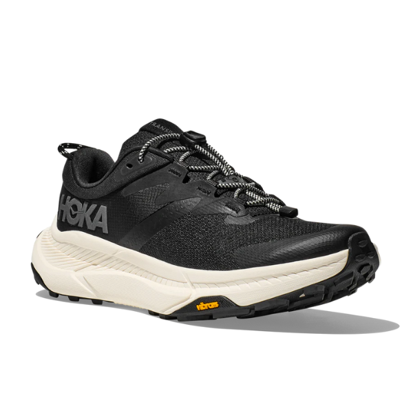 The HOKA Mens Transport Wide Black/White features a black upper, white sole, “HOKA” logo on the side, black and white laces, vegan VIBRAM outsole, shown from an angled front left view against a white background.