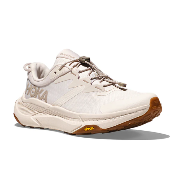 The HOKA Womens Transport Wide Neutral is a beige running shoe with a chunky sole, brown tread, lace-up closure, side HOKA logo, VIBRAM outsole for grip, and is made from vegan materials.