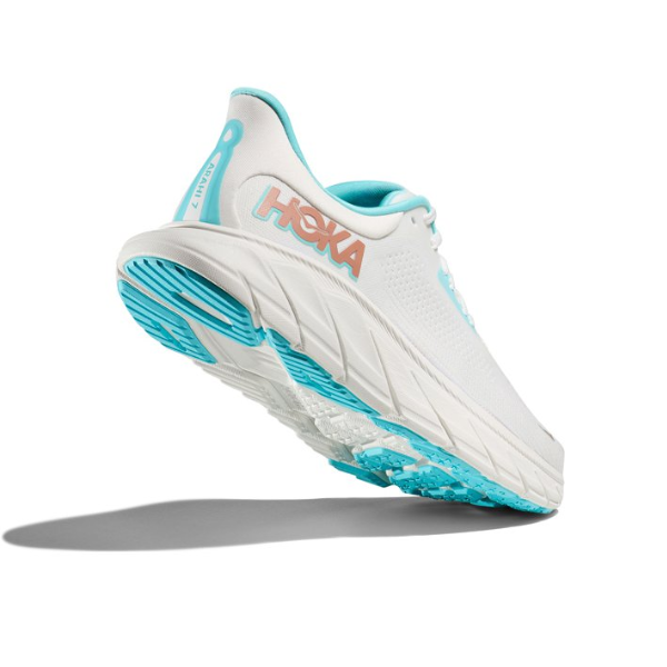 A HOKA Womens Arahi 7 (Medium Width) White/Gold stability shoe is shown from a low rear angle, highlighting its cushioned sole with J-Frame™ technology as it appears to float above a gray shadow on a white background.
