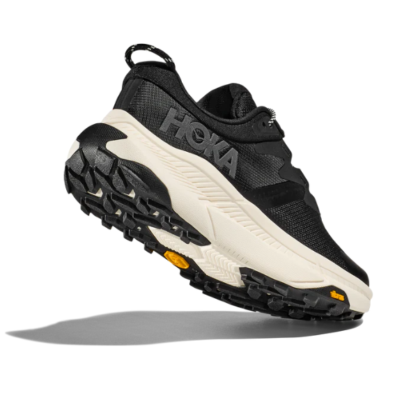 A HOKA Mens Transport Wide Black/White athletic shoe with a rugged VIBRAM outsole and textured tread is displayed at an angle, emphasizing its sole design and casting a shadow on a white background.
