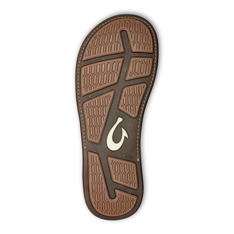 The OLUKAI Mens Tuahine Toffee sandal features a brown sole with textured hexagonal tread, intersecting grip grooves, and Hawaiian-inspired accents, including a central fishhook-shaped logo.
