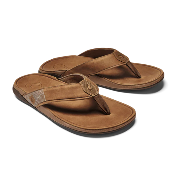 The OLUKAI Mens Tuahine Toffee sandals feature brown leather flip-flop design with cushioned soles and Hawaiian-inspired fabric-accented straps, shown on a white background.