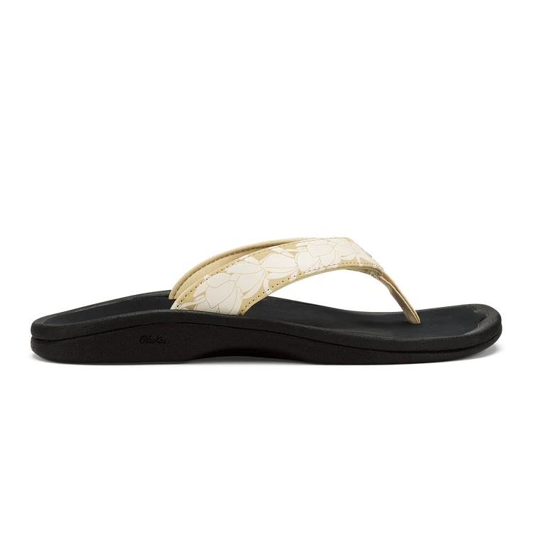 A side view of the OLUKAI Womens Ohana Toasted Coconut sandal featuring a black base, cream floral-patterned strap, water-resistant anatomical footbed, and low heel.