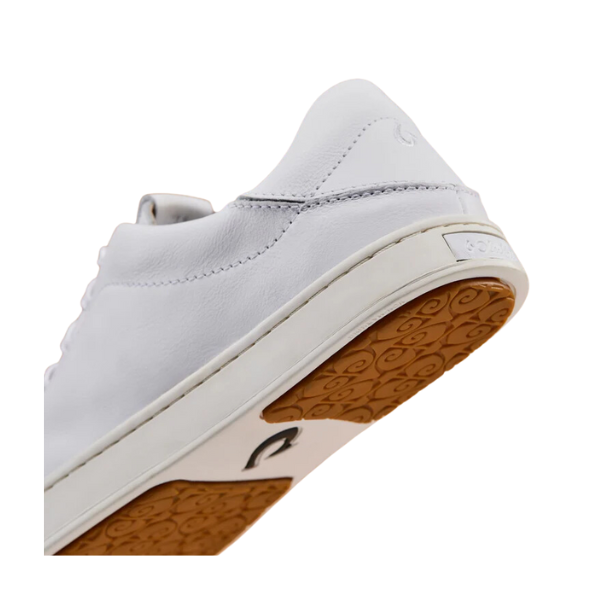 A close-up of the OLUKAI Womens Pehuea Lī ‘Ili Leather White sneaker shows its tan, swirl-patterned rubber outsole, textured details, and a subtle embossed logo on the heel tab—highlighting comfort and support for foot pain.