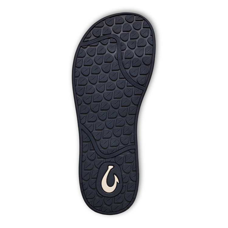 The image shows the navy blue sole of the OLUKAI Womens Southshore Indigo sandal, featuring a textured geometric pattern, a white hook-shaped logo by the heel, and a black outline.