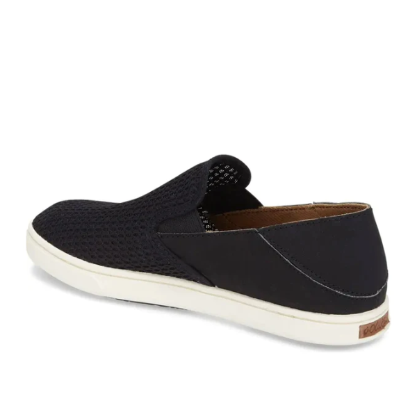 The OluKai Womens Pehuea Black slip-on sneaker features a mesh-textured upper, white sole, anatomical footbed, and a brown back heel accent. The shoe is shown angled to display the side and part of the back.