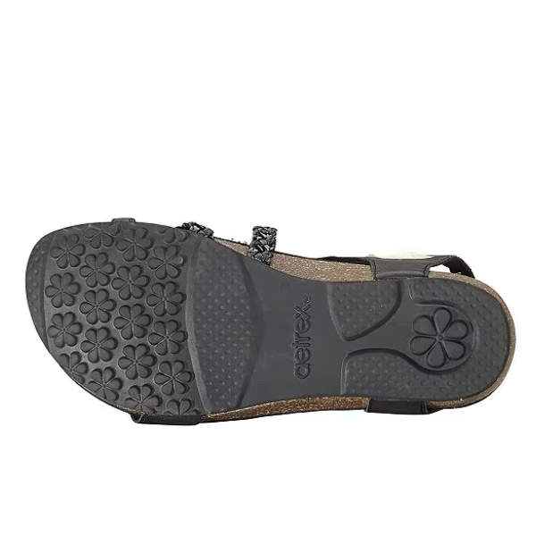 The image shows the bottom view of the Aetrex Womens Jillian Black sandal with a black rubber sole featuring floral patterns for traction and aetrex embossed in the center, plus an orthotic footbed, cork base, and black straps.