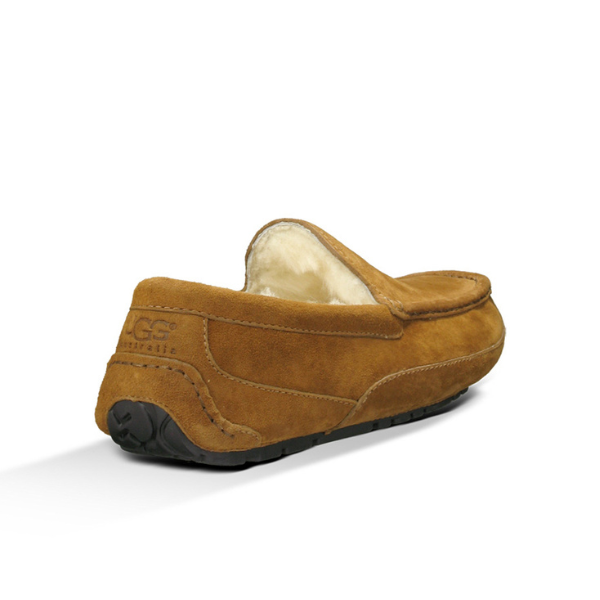 The UGG Mens Ascot Slipper Chestnut is a tan suede loafer with white fleece lining, a black rubber sole for comfort, and the UGG logo embossed on the heel, shown from a rear side angle.