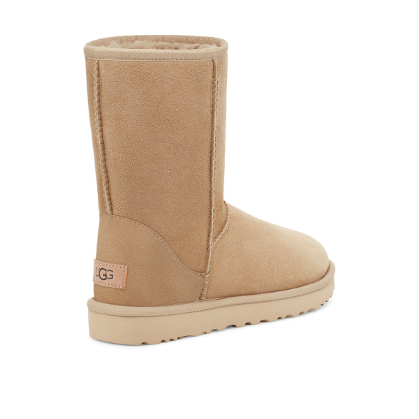The UGG Womens Classic Short II Boot in Mustard Seed is an ankle-high suede boot with a flat sole, visible stitching, and an UGG logo tag above the heel. Its soft interior delivers the signature comfort of sheepskin boots.
