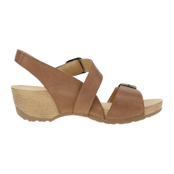 The Dansko Womens Trinity Tan Burnished Nubuck sandal by DANSKO features Wide crisscross straps, an open toe, slingback heel strap, low textured wedge heel, arch support for comfort, and a light wood-like sole with edge stitching.