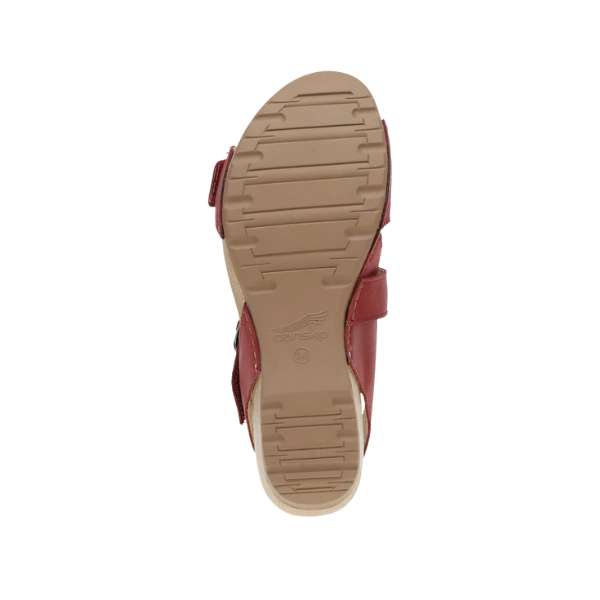 The image shows the bottom view of the Dansko Womens Trinity Cayenne Burnished sandal by DANSKO, featuring a tan, textured rubber sole with horizontal grooves for grip and an embossed Dansko logo near the heel, plus red leather upper straps.