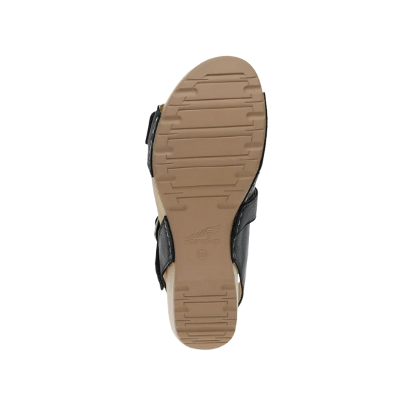 The image shows the bottom view of the Dansko Womens Trinity Black Burnished Nubuck sandal. It features a tan rubber sole with horizontal grooves for grip, black side straps, an embossed DANSKO logo on the midsole, and a contoured footbed for support.