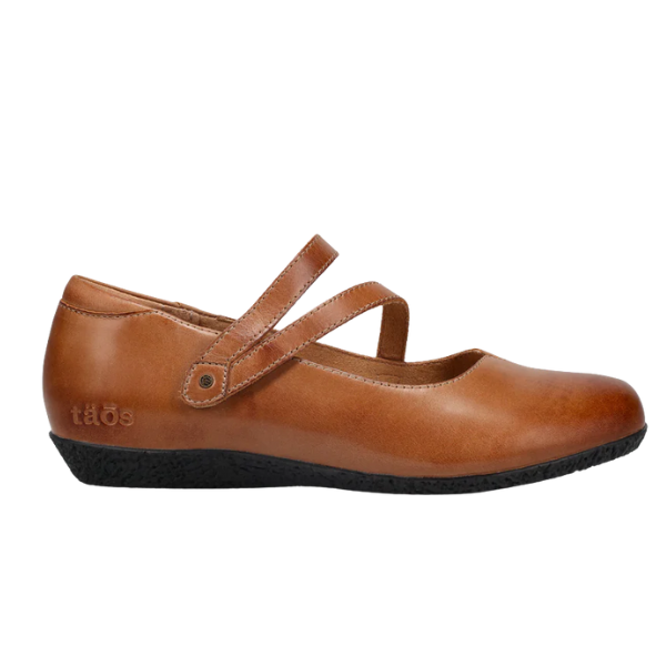 The TAOS Women's Banter Caramel is a brown leather flat with two thin adjustable straps, excellent arch support, a black textured sole, and the TAOS brand embossed near the heel.
