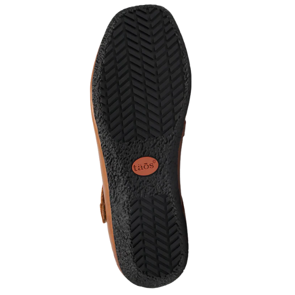 The image displays the sole of the Taos Women's Banter Caramel flat by TAOS, featuring a textured herringbone tread and a circular brown patch with "taös" embossed near the heel, emphasizing quality and arch support.