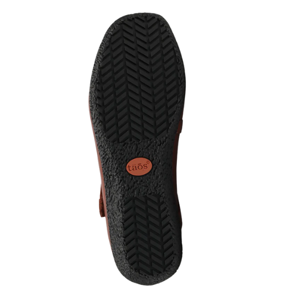 The image shows the black rubber sole of the Taos Women's Banter Cognac by TAOS, featuring a herringbone tread and brown taös logo—crafted for durable comfort and arch support, perfect for those seeking plantar fasciitis relief.