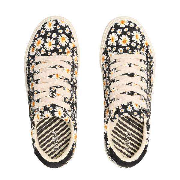 The Taos Women's Z Soul Daisy Black by TAOS is a stylish canvas sneaker with black fabric, white and yellow daisy prints, beige laces, cream soles, arch support for comfort, and insoles featuring bold stripes and the TAOS brand.
