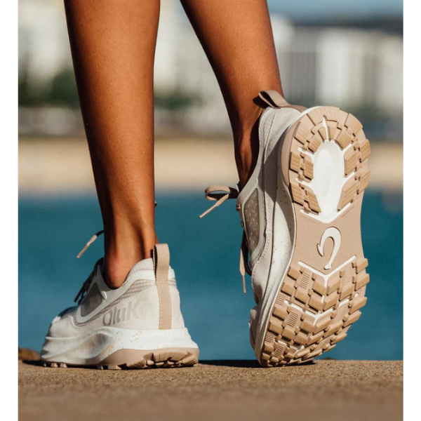 A person wearing OLUKAI Women's Makiki Lava White Sand trail shoes walks on concrete near water, city buildings blurred behind. The distinctive tread showcases their all-terrain grip.
