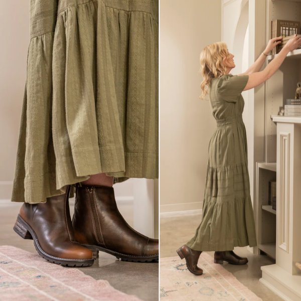 A woman in a long olive-green dress and TAOS Women's Uptown Dark Brown boots stands on a rug, reaching for a book on a tall white bookshelf in a cozy room.