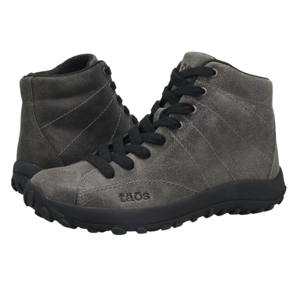 The Taos Women's Trail Mix Charcoal sneakers are gray suede high-tops with black laces and rubber soles, featuring the TAOS brand on the side. They offer a supportive footbed and are styled like boots, shown angled against a white background.