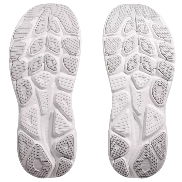Two white athletic shoe soles with textured grips and gray rubber tread highlight the cushioned design of the HOKA Women's Clifton 10 Cosmic Grey, featuring raised HOKA branding.