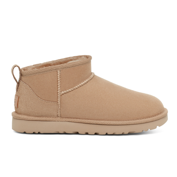 The UGG Womens Classic Ultra Mini Sand is a tan suede ankle boot with plush lining, rounded toe, back pull tab, and flat rubber sole—versatile footwear by UGG shown in a side profile view.