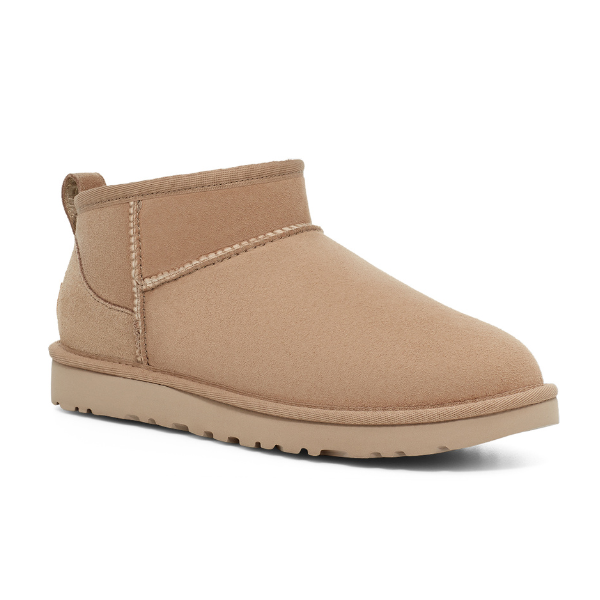 The UGG Womens Classic Ultra Mini Sand is a versatile suede ankle boot with a rounded toe, visible stitching, heel pull tab, and flat rubber sole inspired by the brands iconic design.