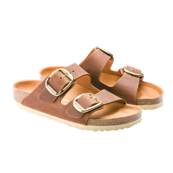 The Birkenstock Womens Arizona Big Buckle Cognac sandals by BIRKENSTOCK feature two large gold buckles and cork soles, crafted in oiled brown nubuck leather and displayed on a white background.