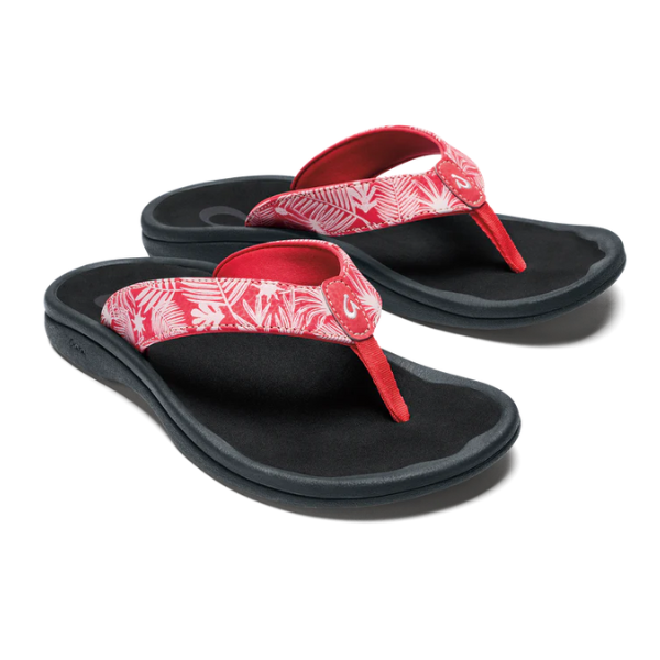 The OLUKAI Womens Ohana sandals in Coral/Lehua Flower/Onyx feature thick red straps with a white tropical leaf pattern and black soles. These water-resistant flip-flops offer an anatomical footbed and cushioned comfort for everyday wear.