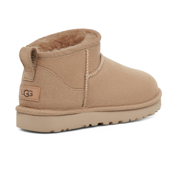 The UGG Womens Classic Ultra Mini Sand boot features tan suede, plush lining, visible stitching, a heel pull tab, tan rubber sole, and the UGG logo on a tag at the back.