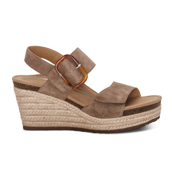 The Aetrex Womens Ashley Espadrille Wedge Taupe by AETREX WORLDWide INC is a taupe suede sandal with a thick ankle strap, large brown buckle, open toe, woven espadrille platform heel, and a memory foam footbed for comfort.
